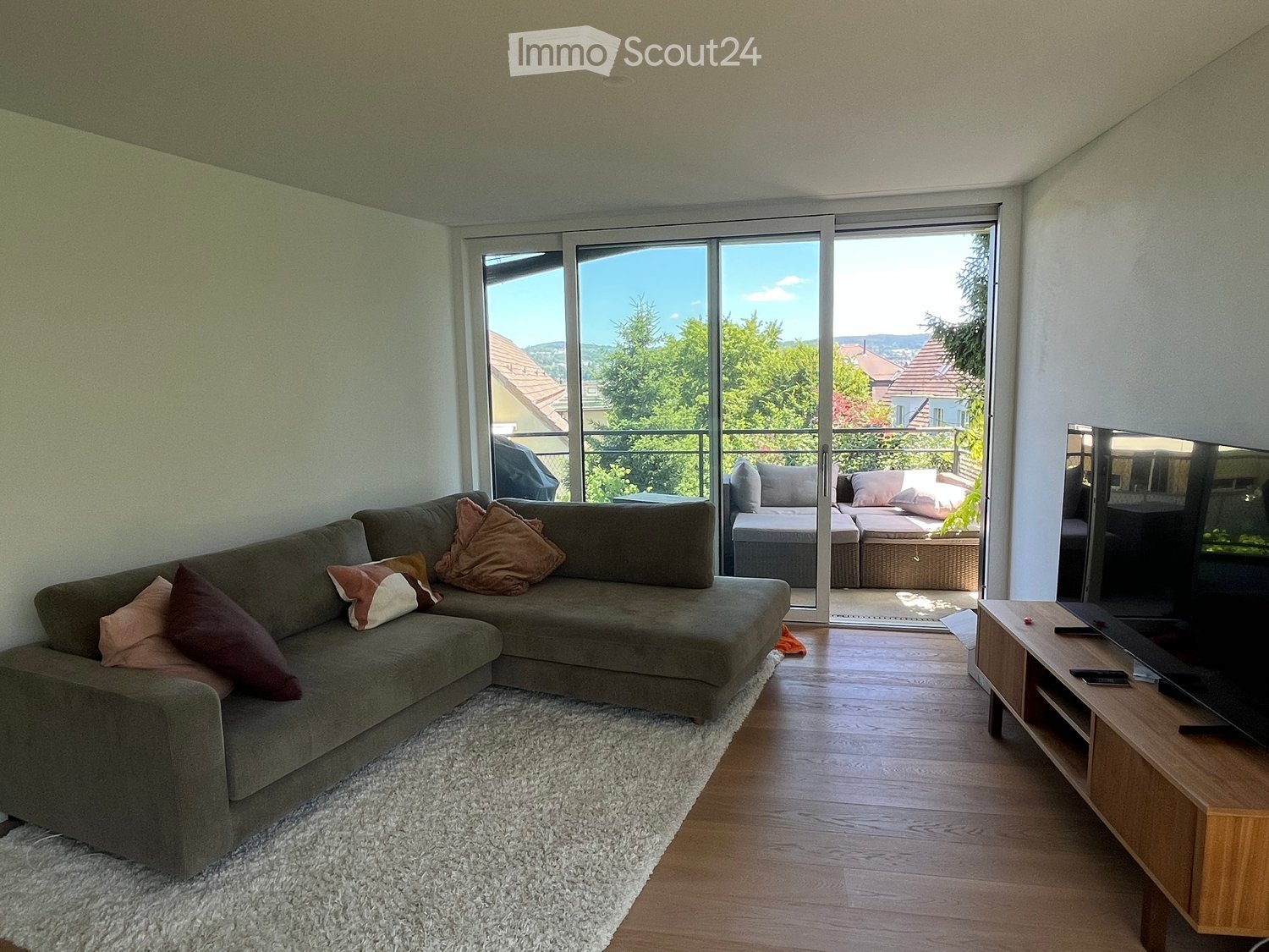 Modern living room with a grey sectional sofa, multiple throw pillows, a flat-screen TV on a wooden stand, and a view of greenery through large glass doors leading to a balcony.
