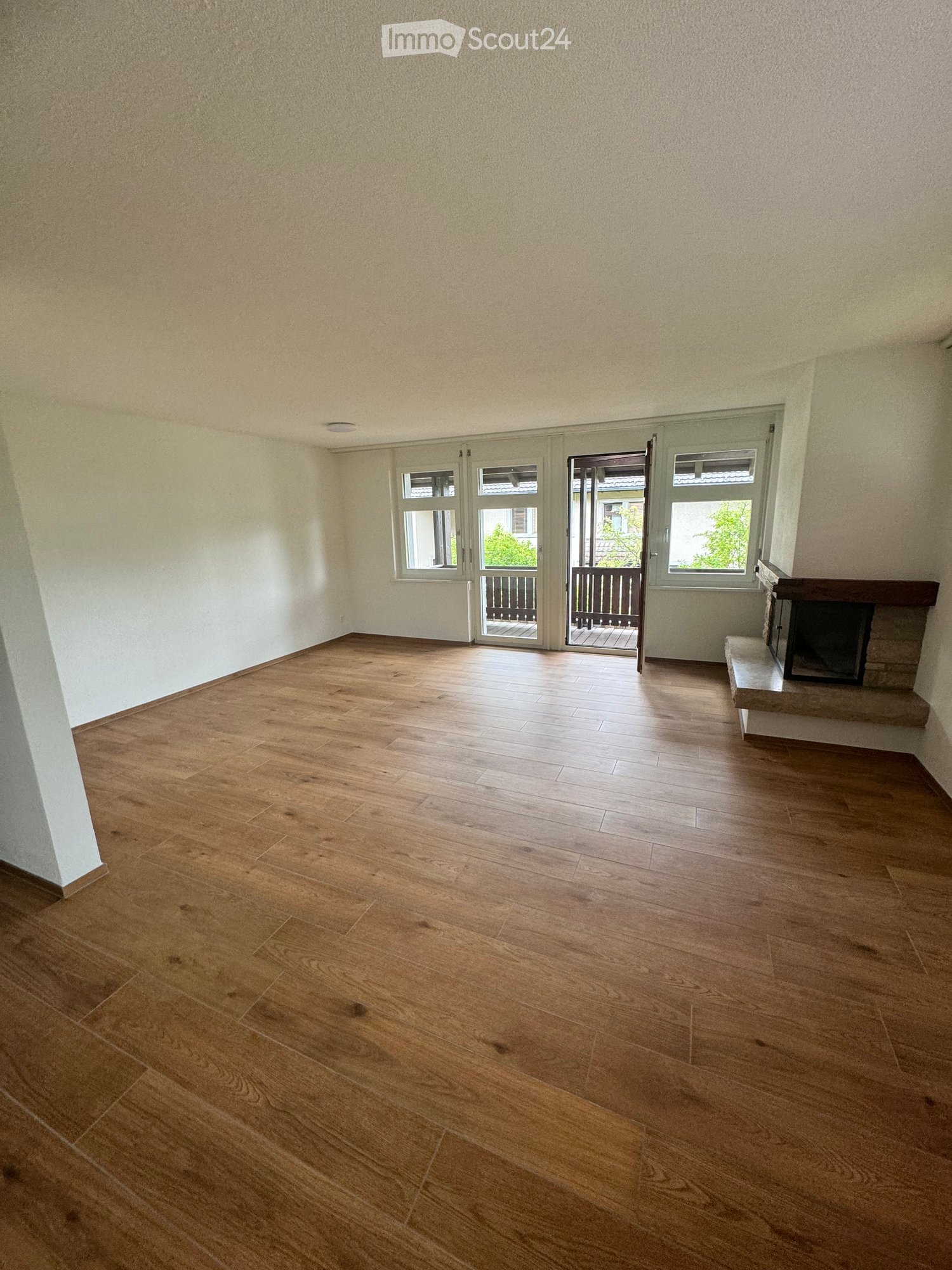 Empty room with wooden flooring, white walls, and a fireplace on the right side. There are large windows and a door leading to a balcony.