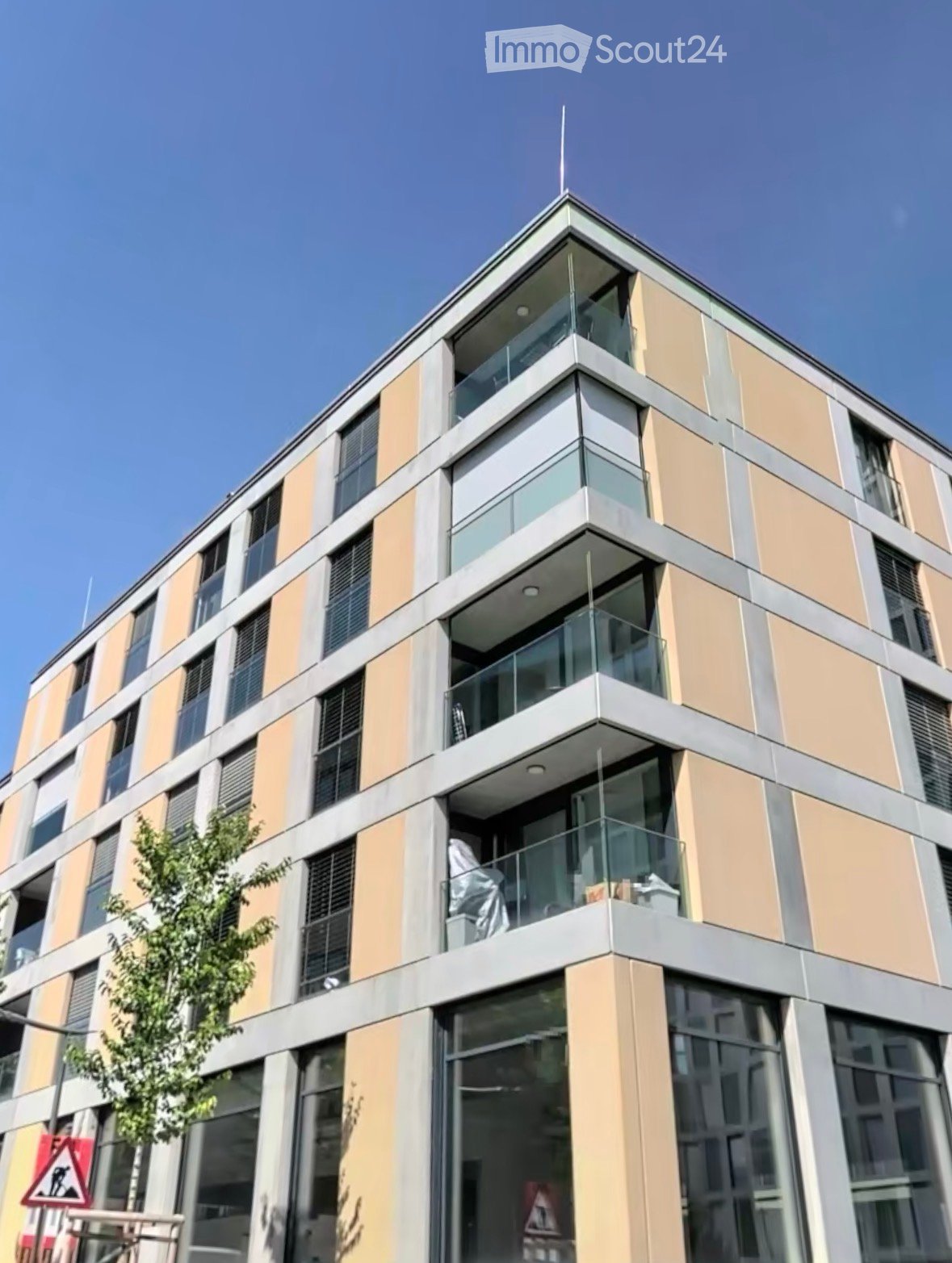 Modern multi-story building with beige and grey facade, large windows, and balconies with glass railings.