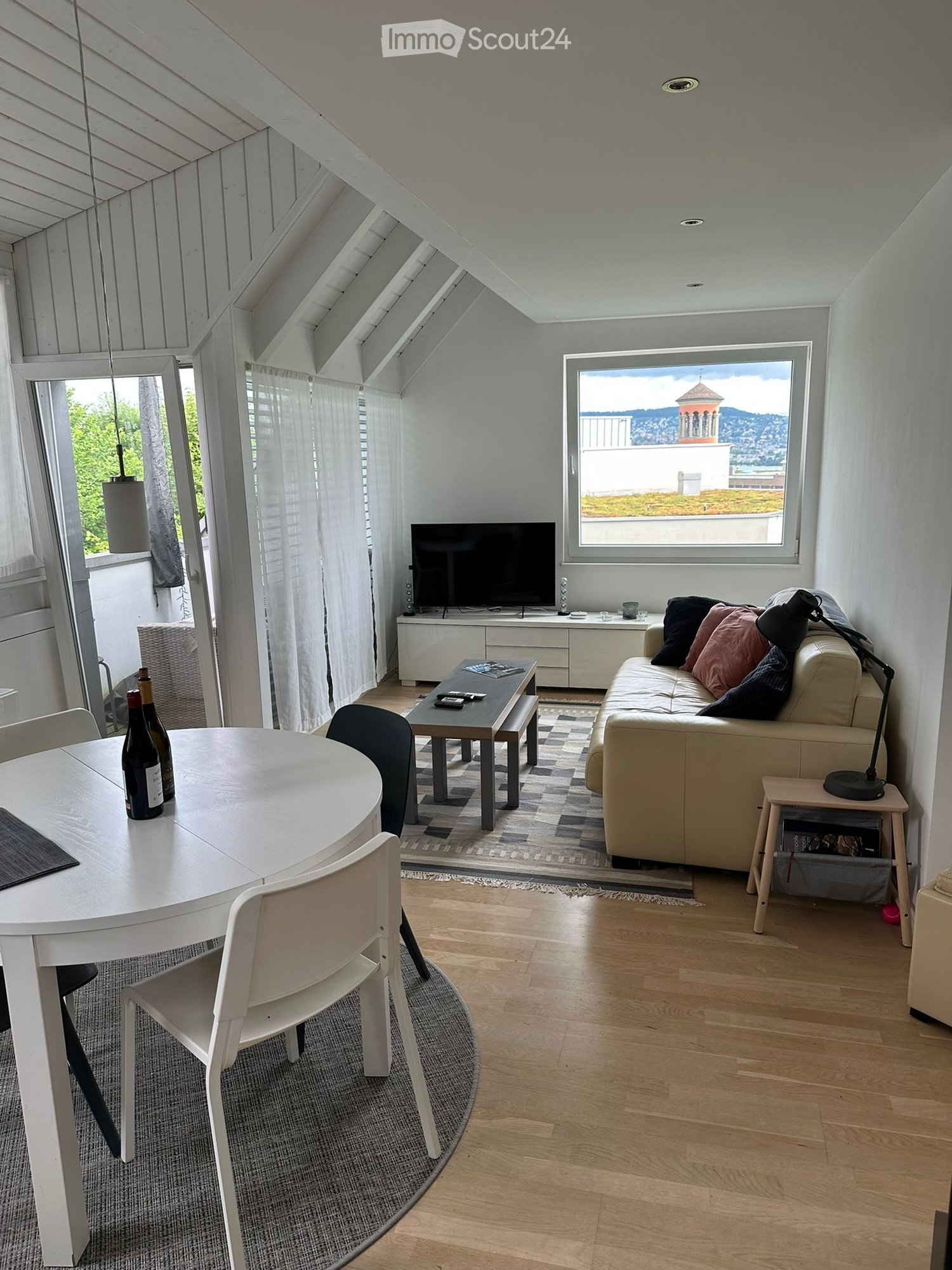 Bright living and dining area with wooden floors, large windows offering a scenic view, a sofa with cushions, a coffee table, a dining table with chairs, and a television on a stand.