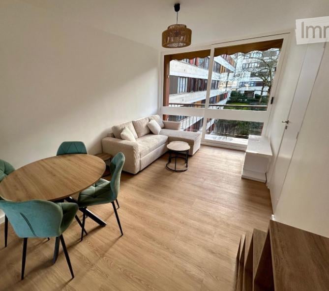 Bright living room with large windows and balcony access, featuring a light-colored sofa, coffee table, dining table with chairs, and a hanging floor lamp.