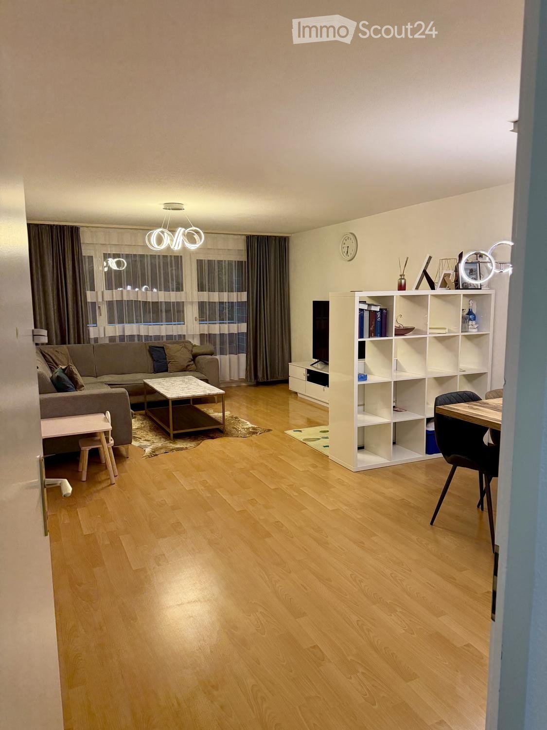 Modern living room with wooden flooring, large windows, a grey sofa, a coffee table, a shelf with books and decorative items, a dining table with chairs, and a small stool near the sofa.