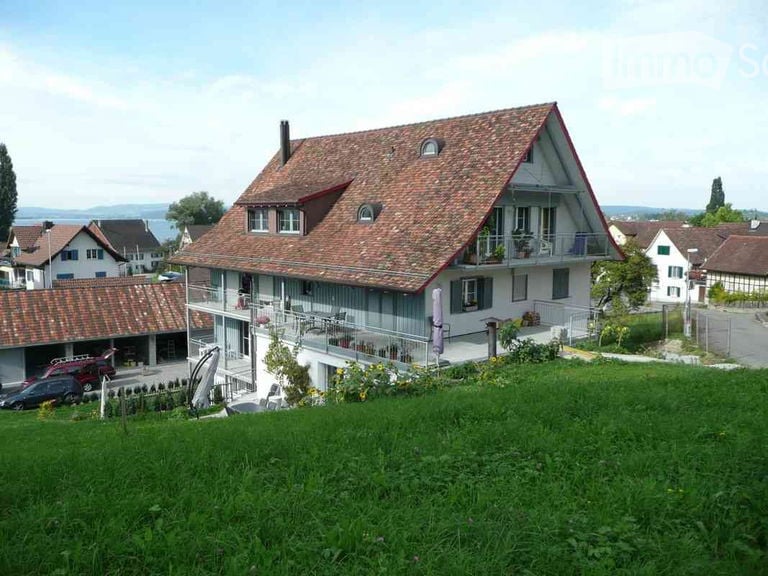 Two-story house with a red-tiled roof, white walls, and a balcony overlooking a green lawn. Garage and parking area visible in the foreground.