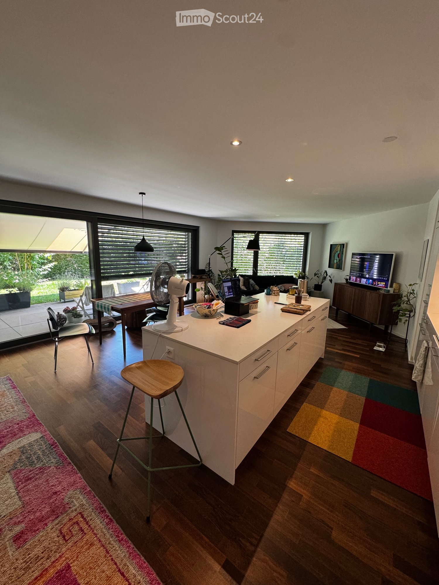 Modern open-plan living and dining area with a white kitchen island, wooden flooring, colorful rugs, and large windows with blinds. Includes a fan, laptop, TV, and various seating options.