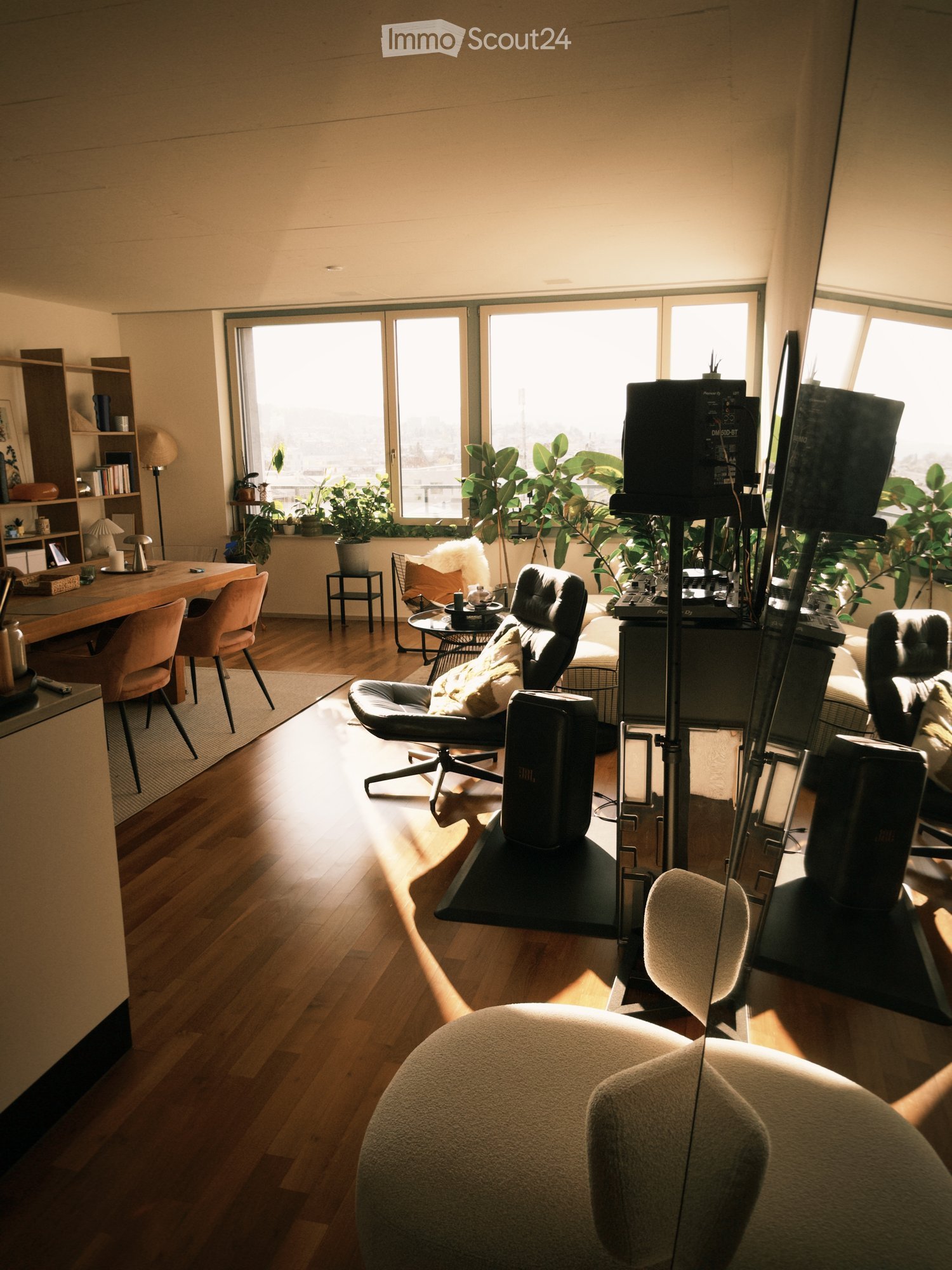 Bright, modern living room with wooden flooring, large windows, and a mix of seating including a sofa, chairs, and a round ottoman. There is a coffee table, a shelf with books and decor, and several plants near the windows.