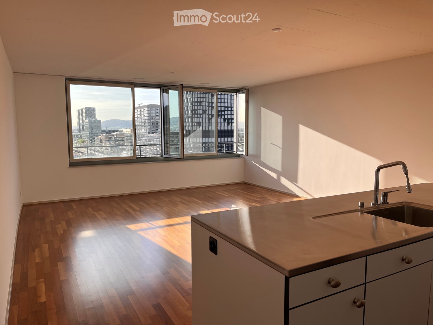 Modern kitchen with wooden flooring, large windows offering a city view, and a stainless steel sink with a modern faucet.