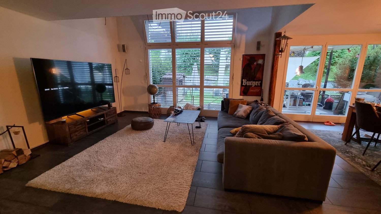 Modern living room with large windows, a grey sectional sofa, coffee table, ottoman, TV, floor lamp, globe, and a dining area visible through the open sliding doors.