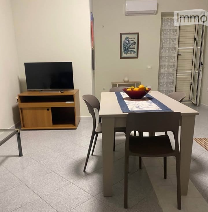 A modern dining area with a white table, four chairs, a TV on a wooden stand, and a bowl of fruit on the table. The room has tiled flooring and an air conditioning unit on the wall.