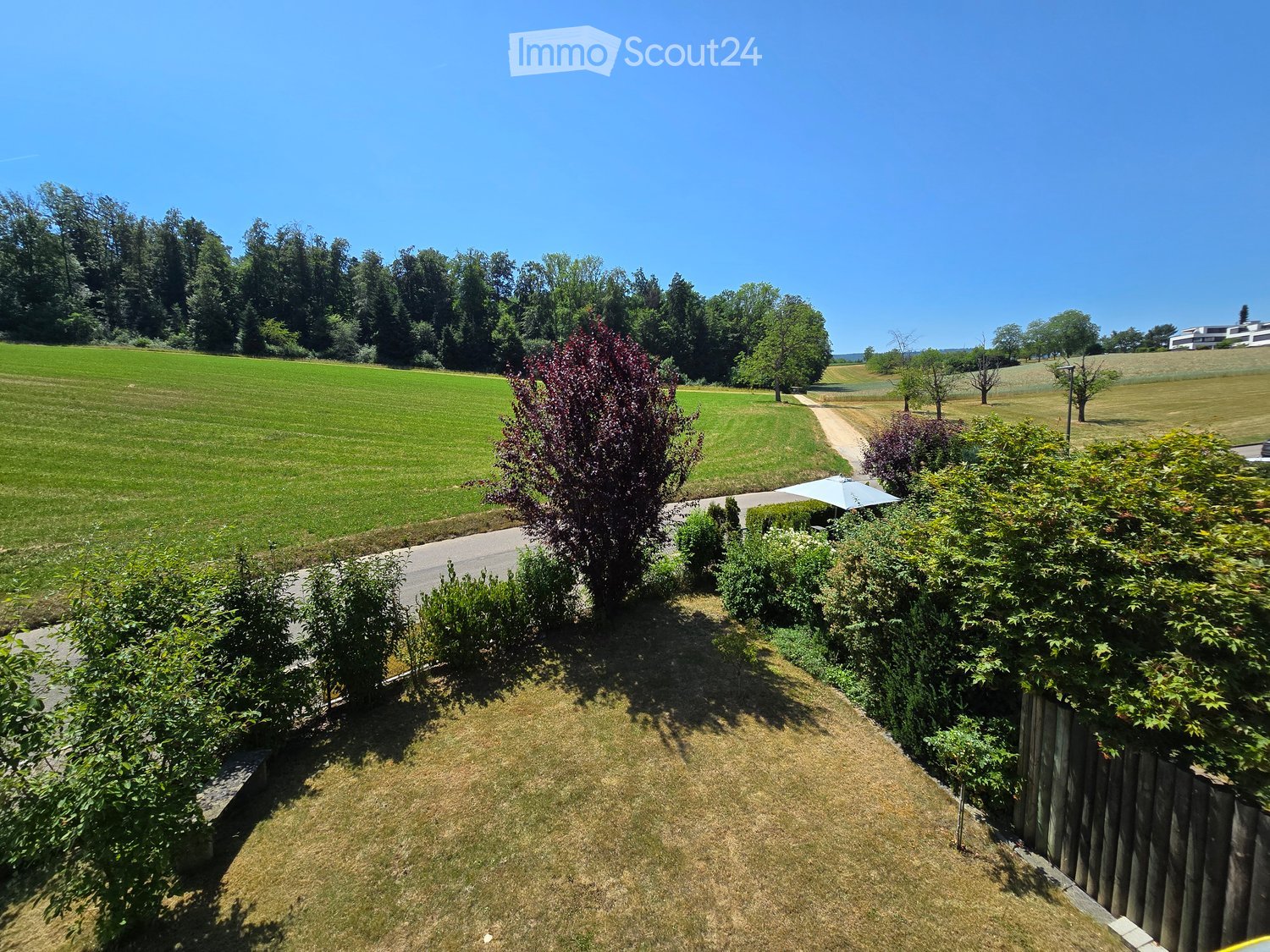 A well-maintained garden with a wooden fence, various shrubs, and trees, leading to a grassy field and a road.