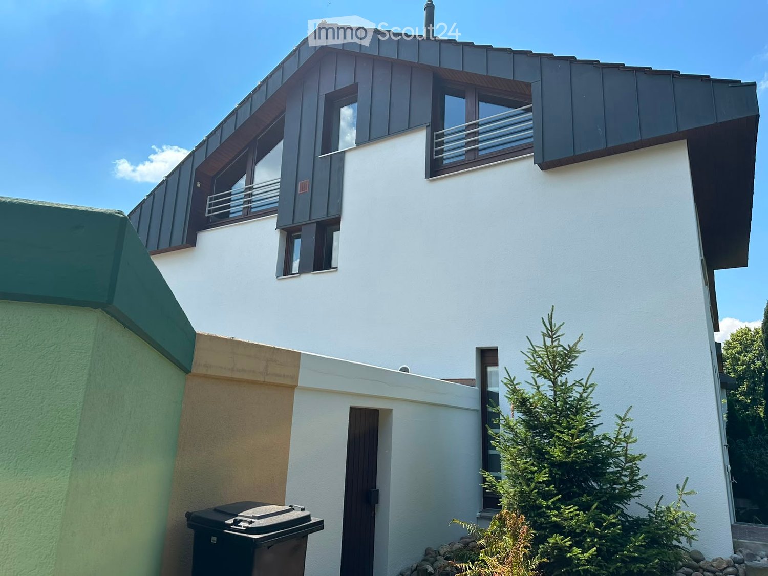 Modern two-story house with white walls, dark roof, and large windows. Features a balcony on the upper floor and a small garden area with a pine tree.