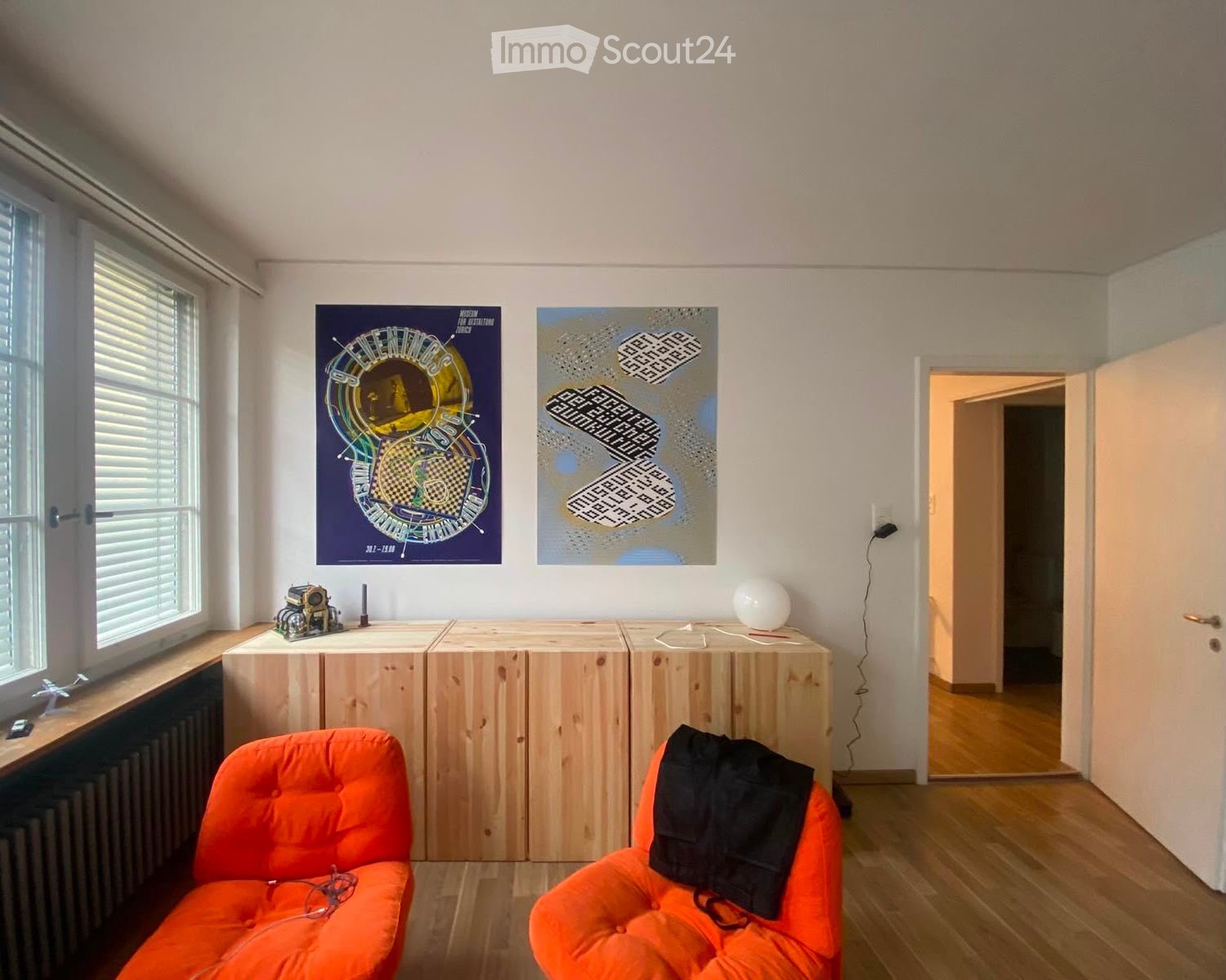 Bright living room with wooden flooring, two orange bean bag chairs, a small table with a lamp, and two posters on the wall. A doorway leads to another room.