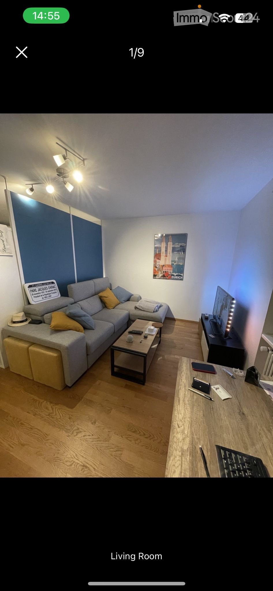 Modern living room with a grey sectional sofa, wooden coffee table, TV stand, and a small desk with a laptop and keyboard. The room features wooden flooring, a blue accent wall, and decorative artwork.