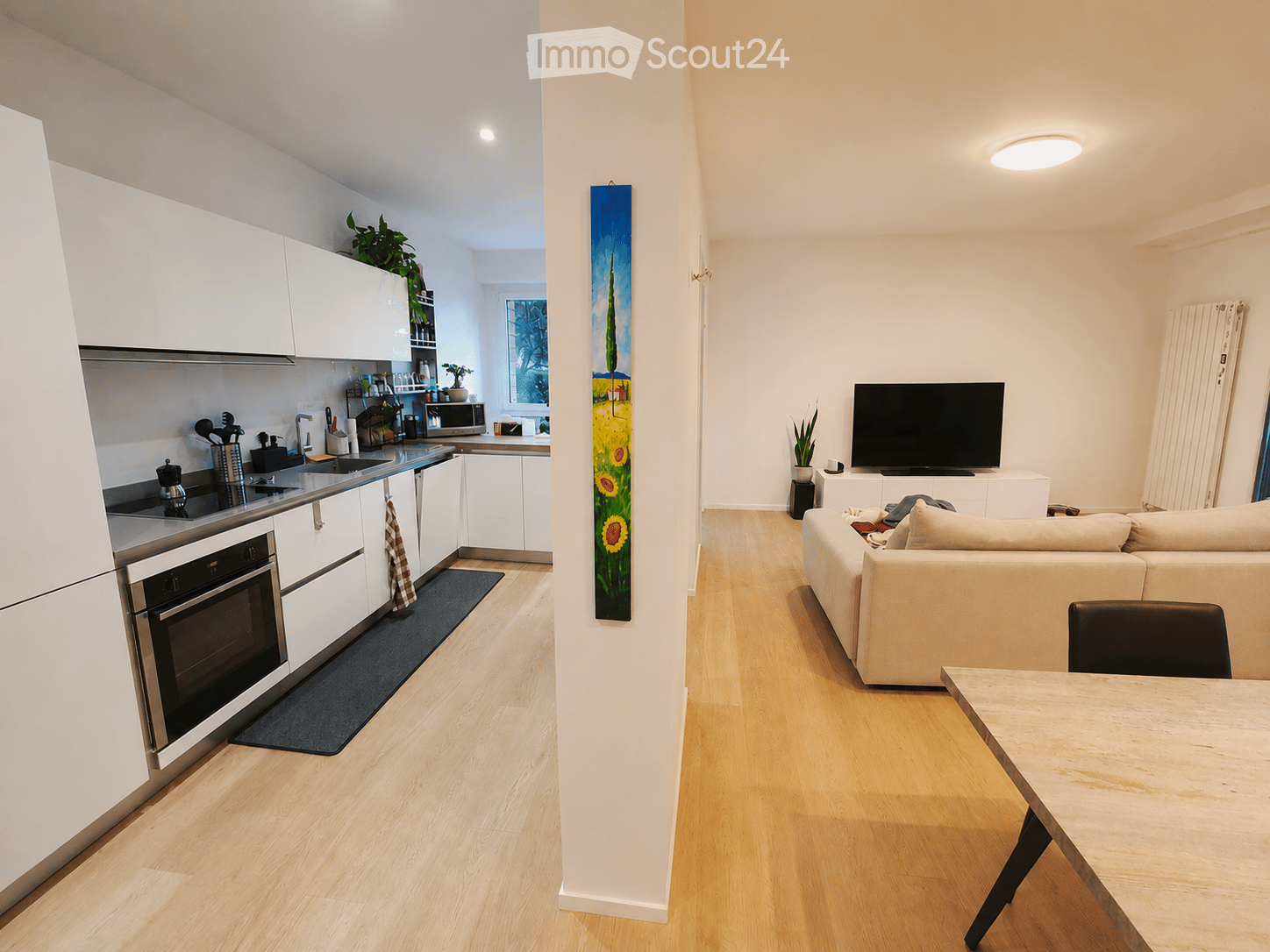 Modern open-plan living and dining area with a kitchen. Features a light-colored sofa, a wooden dining table with chairs, a flat-screen TV, and a sleek white kitchen with stainless steel appliances.