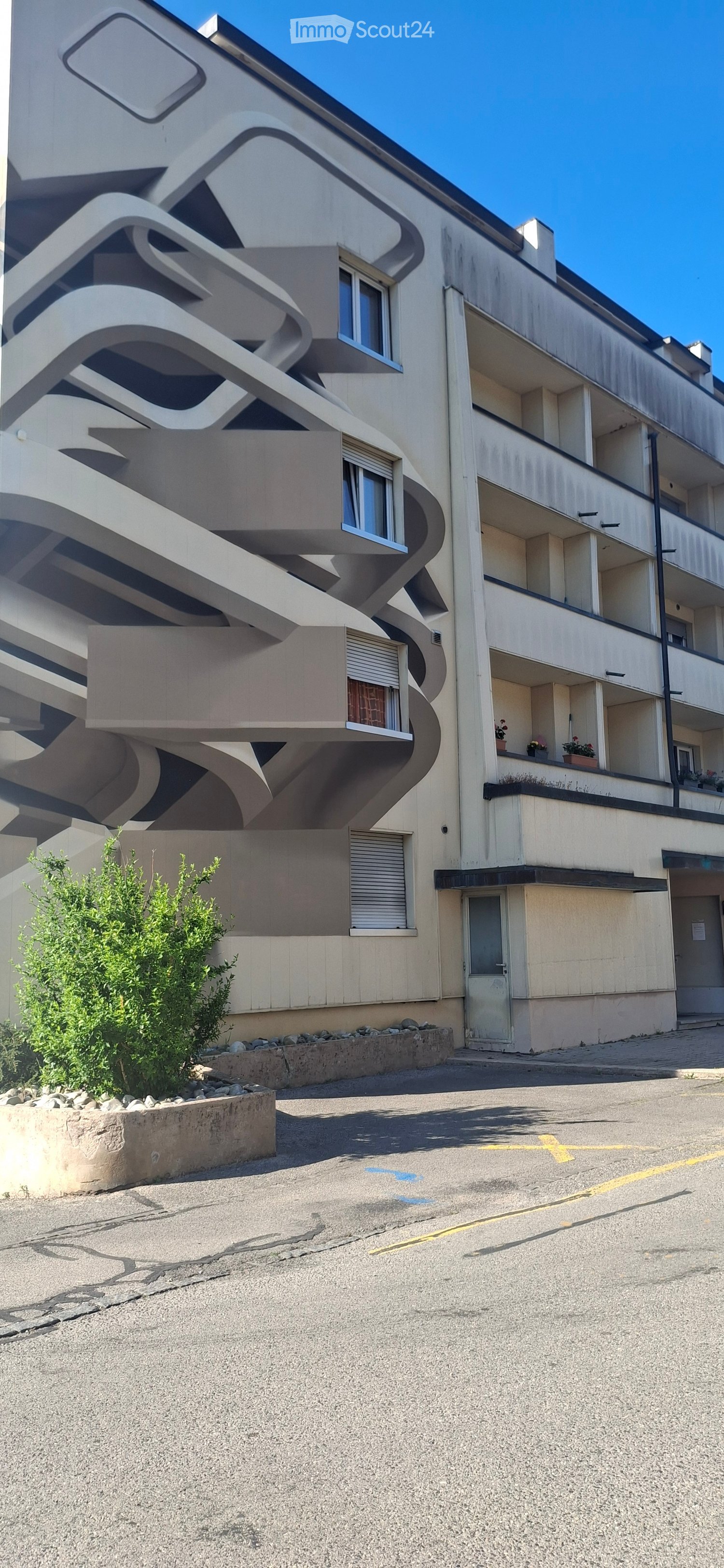 Modern multi-story building with curved balconies and large windows, featuring a small garden area in front.