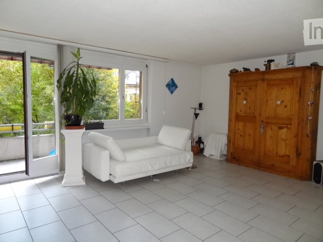 Bright living room with white tiled flooring, large windows, a white sofa, a wooden cabinet, and a potted plant near the balcony entrance.