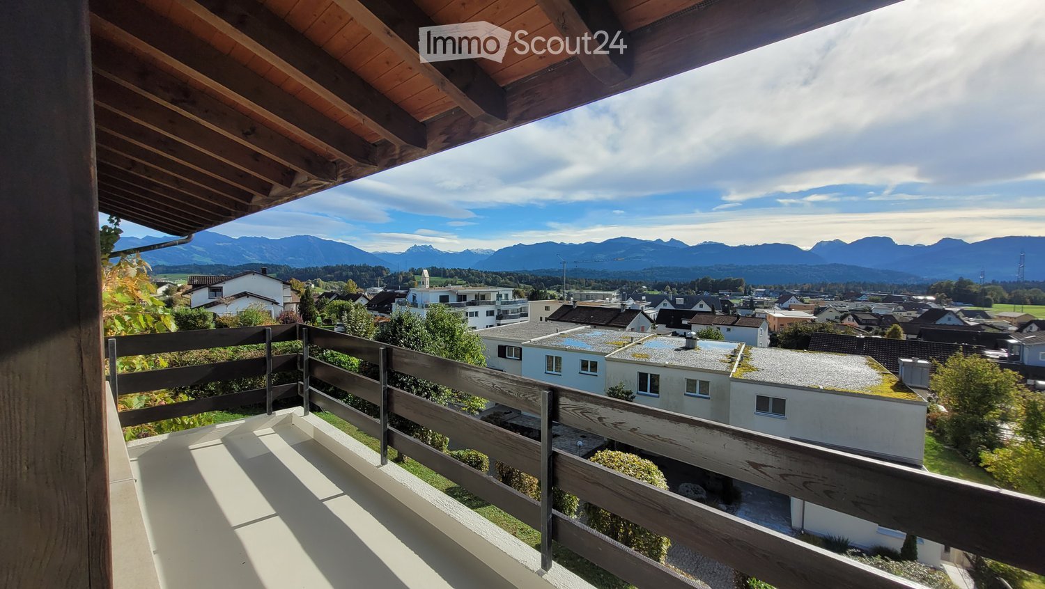Balcony with wooden railing, overlooking a town with houses and distant mountains under a partly cloudy sky.