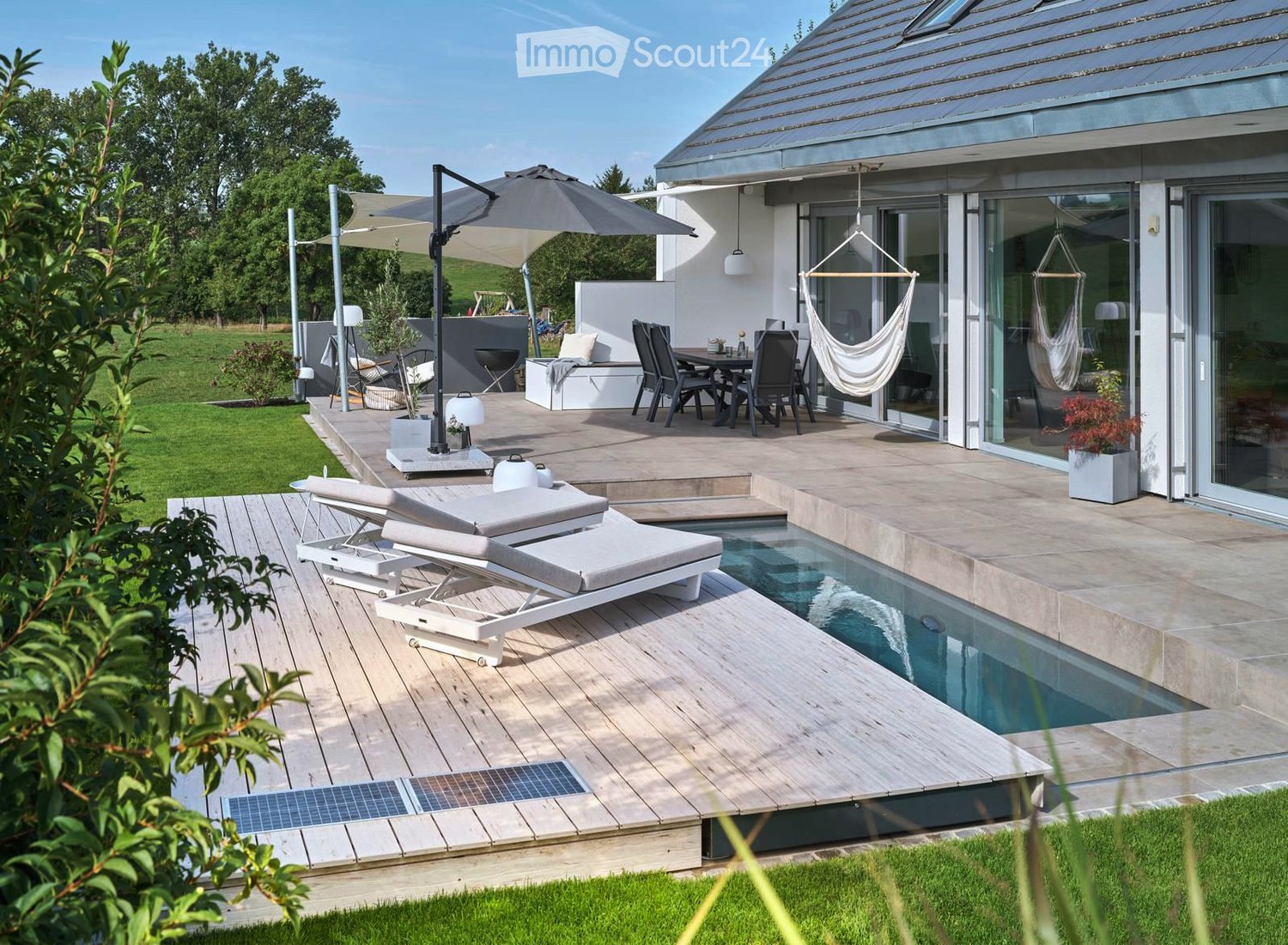 Modern outdoor patio with wooden decking, swimming pool, lounge chairs, dining set, hammock, umbrella, and surrounding greenery.
