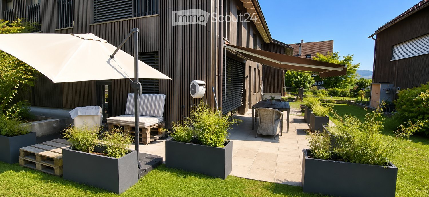 Outdoor patio area with modern furniture, including a table, chairs, and lounge seating, surrounded by potted plants and greenery. Features a large umbrella and a covered seating area.