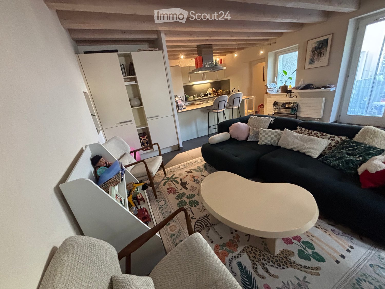 A modern living room with a dark sectional sofa, a round coffee table, and a patterned rug. The room features a kitchen area with a white island, bar stools, and modern cabinets. A cozy armchair and a small shelf with toys are also visible.