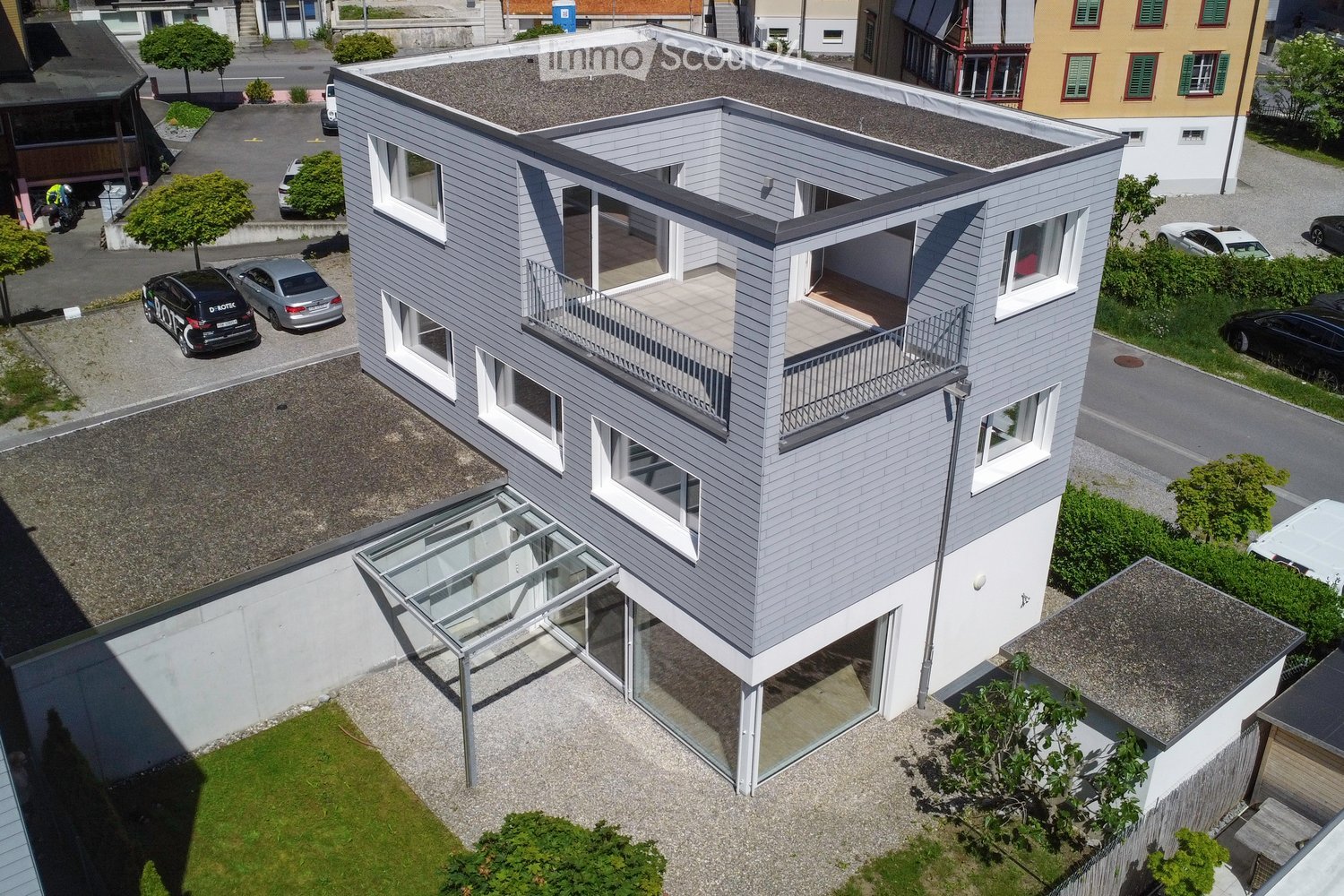 Modern two-story house with grey siding, white trim, and a flat roof. Features include large windows, a balcony on the upper level, and a covered entrance area with glass panels.