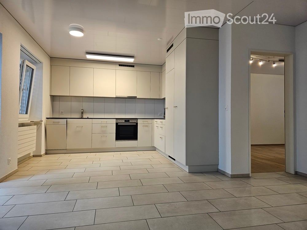 Modern, minimalist kitchen with white cabinets, a built-in oven, and a dishwasher. The space features tiled flooring and ample natural light from a window on the left.