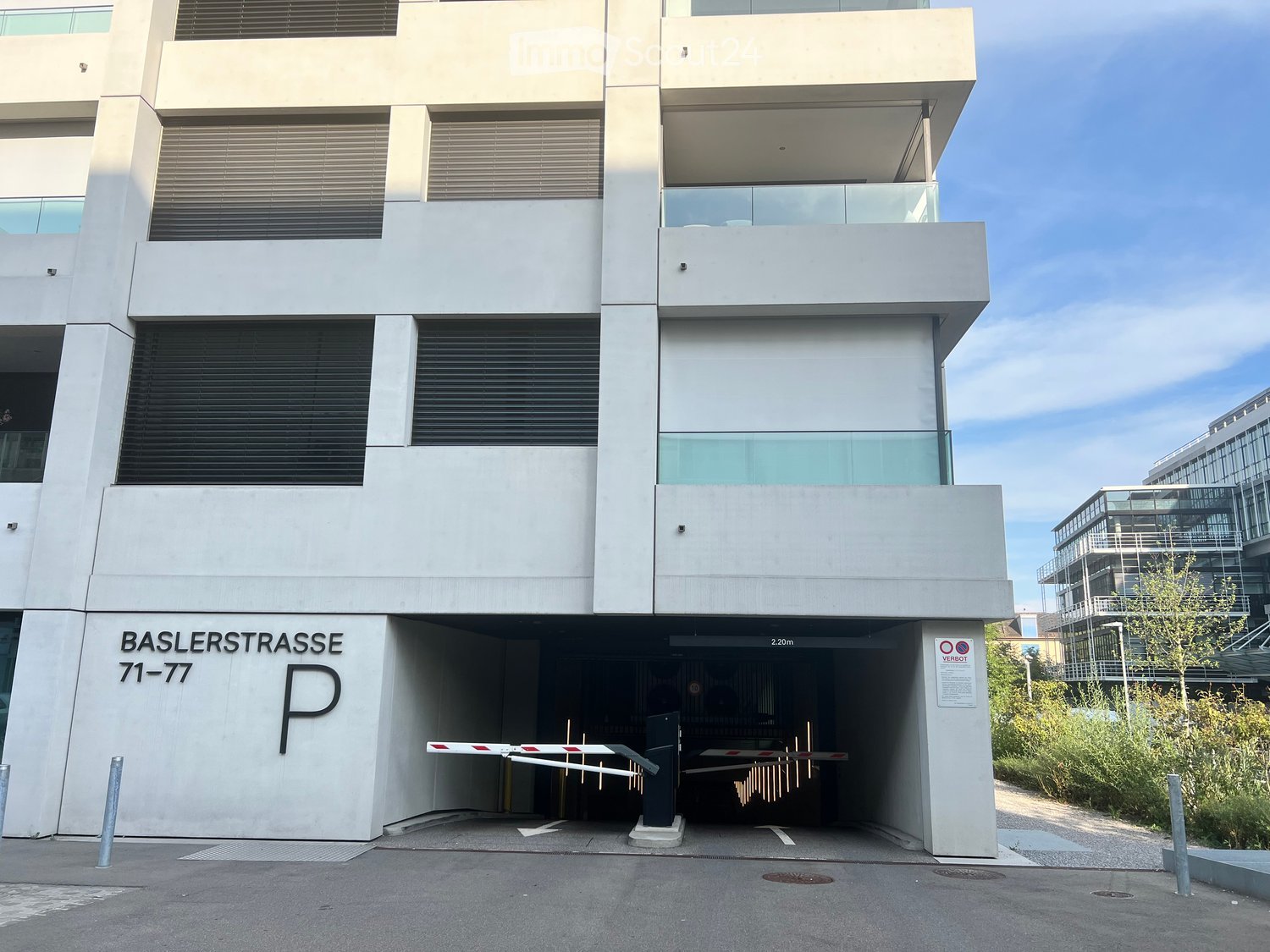 Modern building entrance with a parking area, labeled 'BASLERSTRASSE 71-77', featuring a gate and a clear view of the surrounding area.