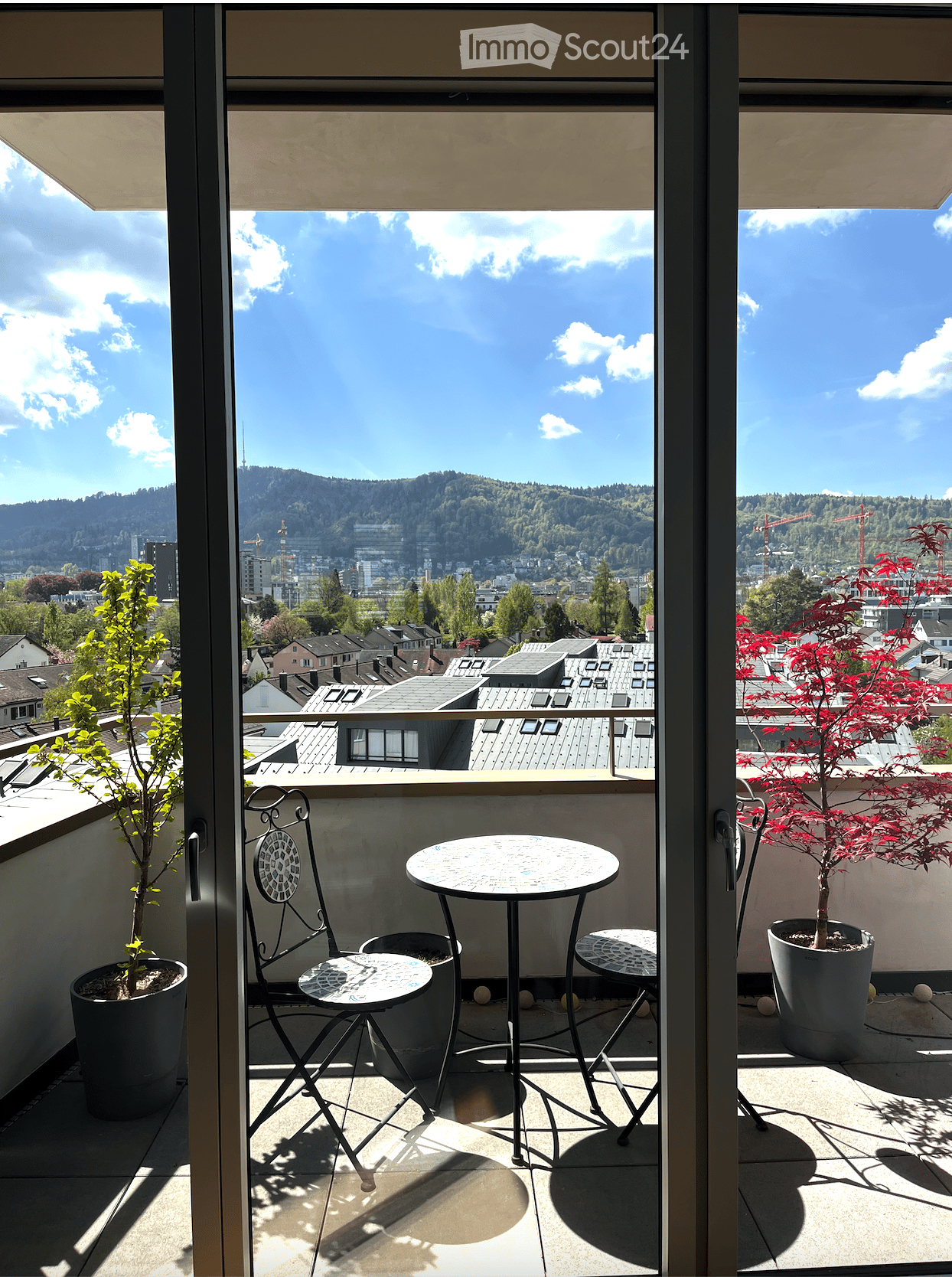 Balcony with a view of the city and mountains, featuring a small round table and two chairs, along with potted plants.