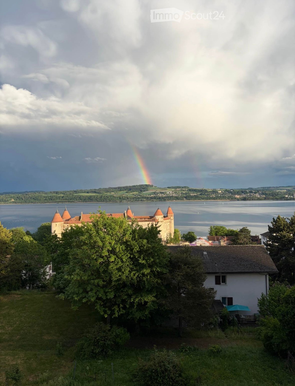 A two-story house with a tiled roof, surrounded by trees and greenery, with a view of a lake and a distant castle in the background under a partly cloudy sky with a rainbow visible.