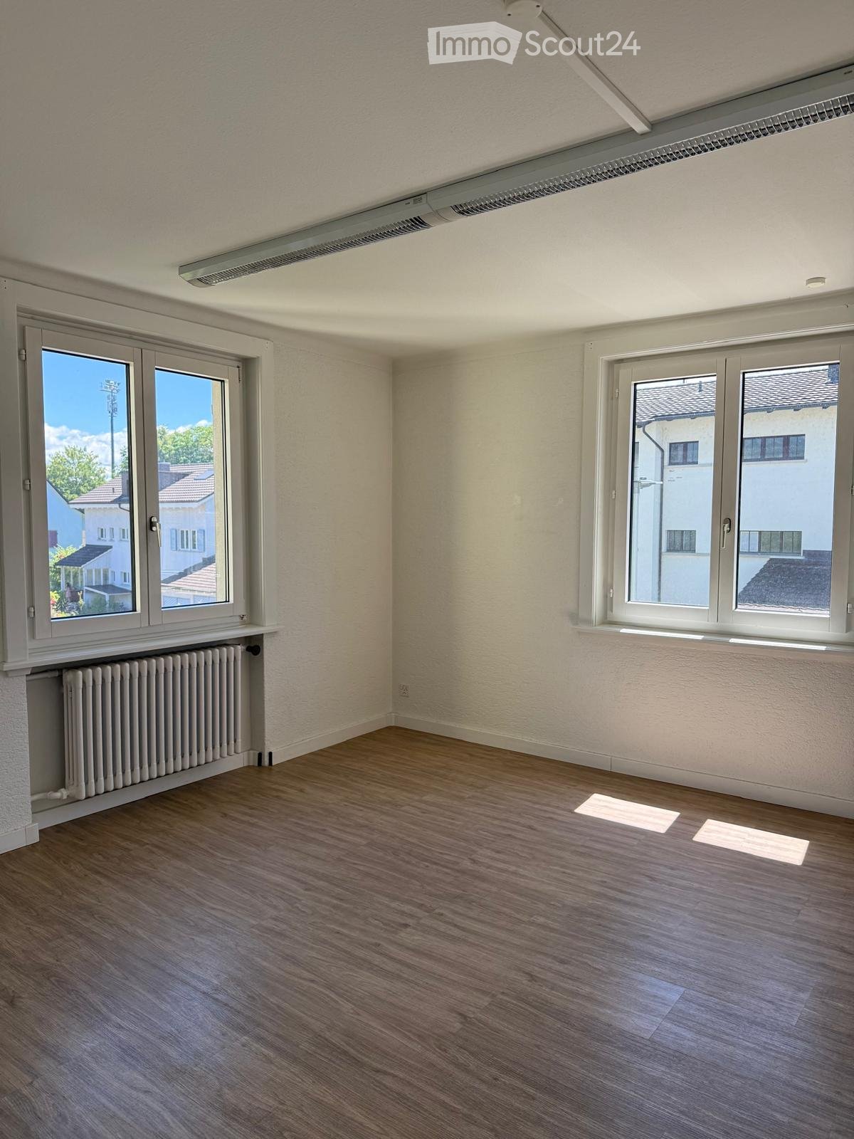 Empty room with wooden flooring, white walls, large windows, radiator, and visible electrical outlets.