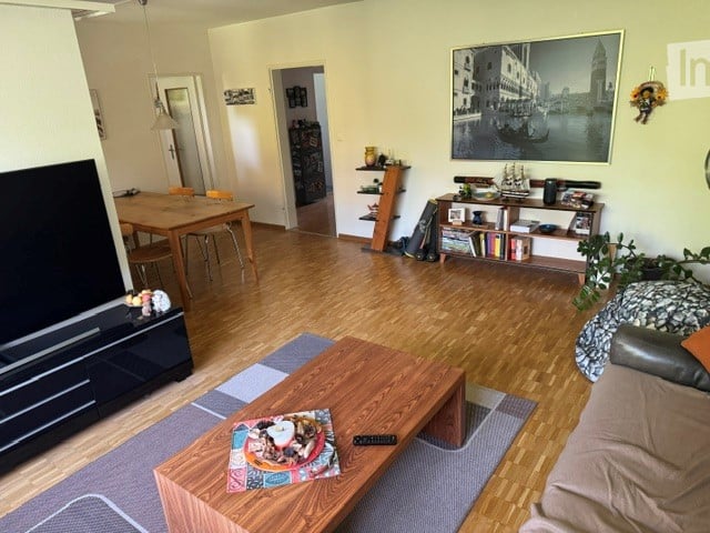 A well-lit living room with hardwood flooring, a sofa, coffee table, dining area with a table and chairs, bookshelf, and a television. The room features a large painting on the wall and a small plant in the corner.