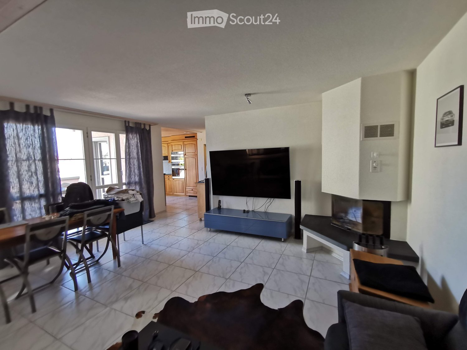 A modern living room with a sofa, coffee table, dining table, chairs, television, and fireplace. The room features large windows with curtains and a view of the kitchen area.