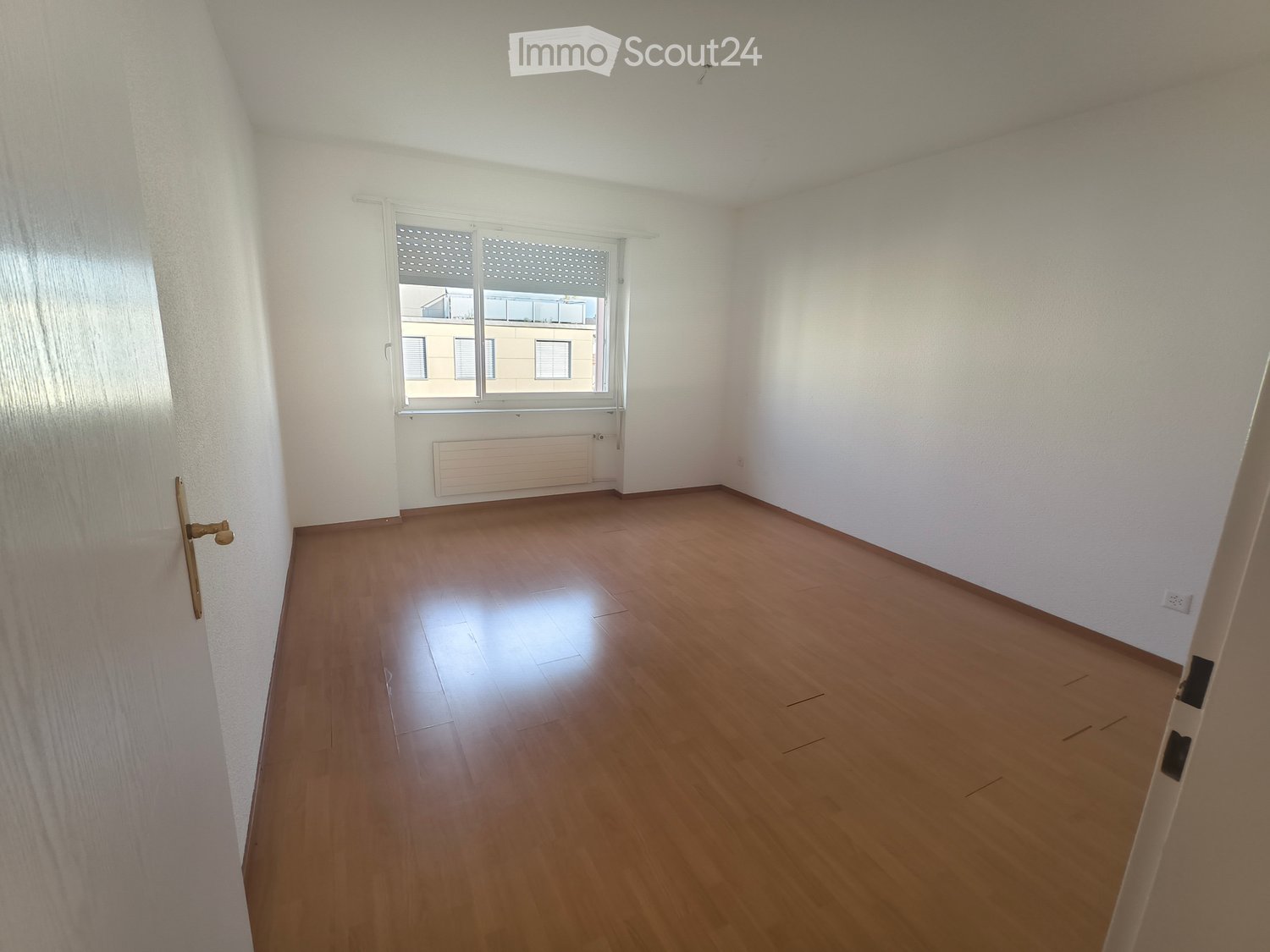Empty room with wooden flooring, white walls, and a large window with blinds. A radiator is visible below the window, and a door is on the left side.