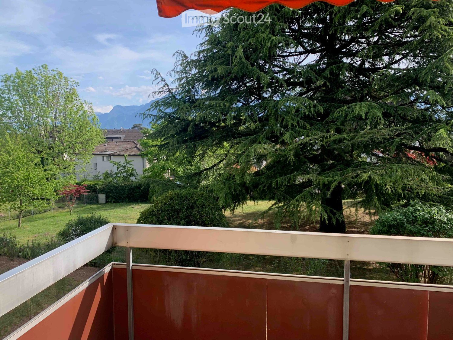 Balcony with wooden railing, overlooking a green garden and trees, with a distant view of mountains.