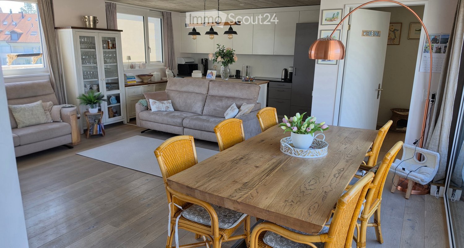 Bright living and dining area with wooden flooring, a large sofa, a dining table with chairs, a floor lamp, and a cabinet with glass doors.