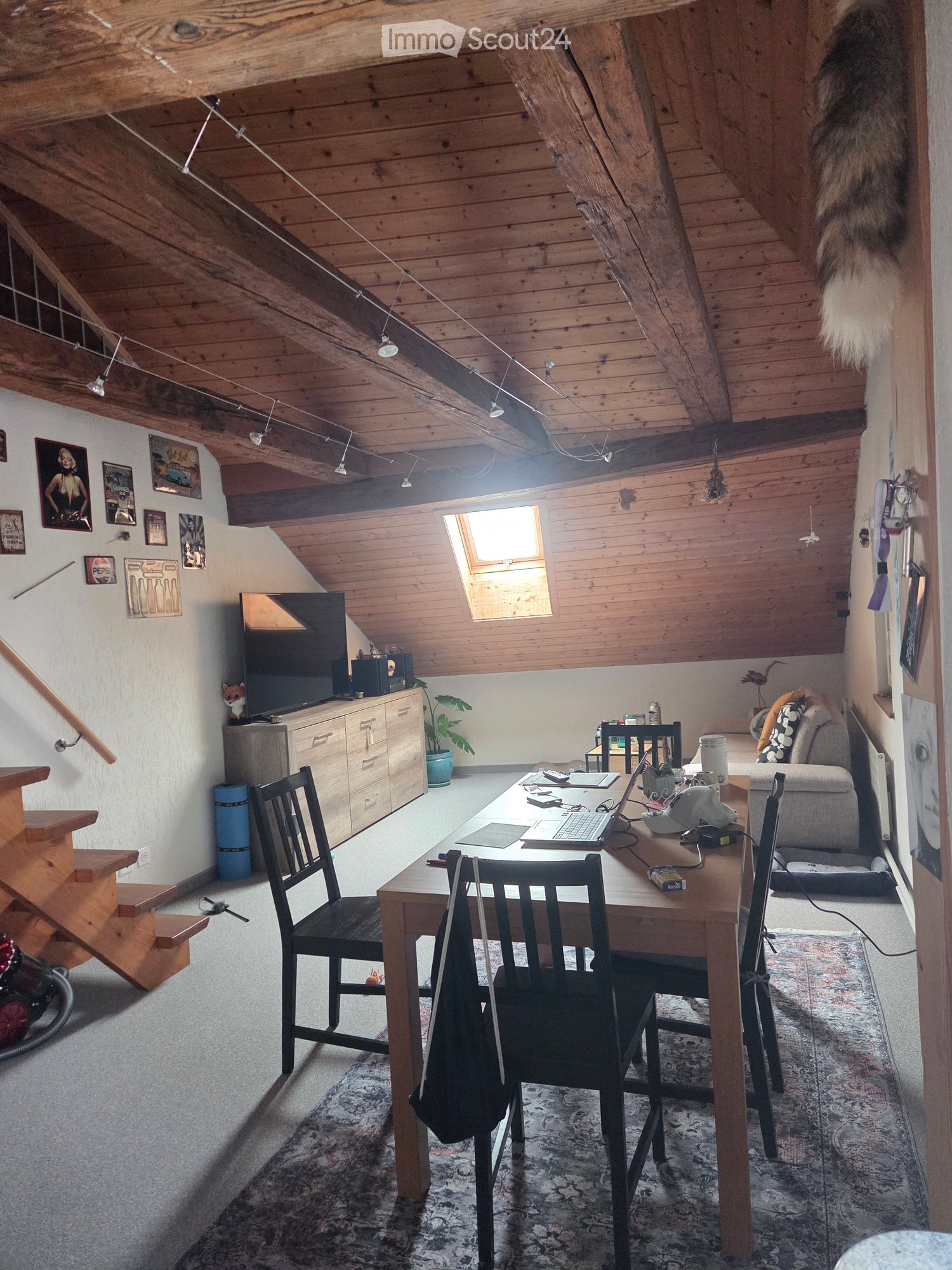 A cozy attic room with wooden beams, a large skylight, a wooden table with chairs, a laptop, a sofa, a television, and a plant. The room features exposed wooden beams and a staircase leading up.