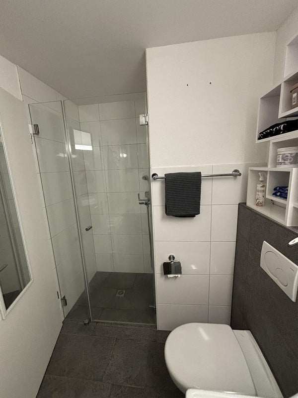 Modern bathroom with a glass-enclosed shower, white tiled walls, a toilet, a towel rack with a black towel, and a shelf with toiletries.