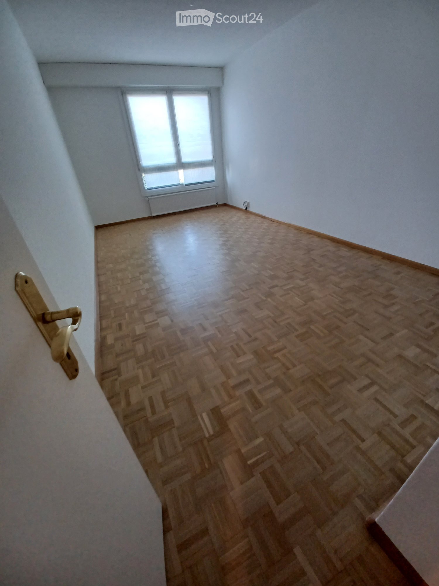 Empty room with wooden herringbone flooring, white walls, and a single window with radiator below it.