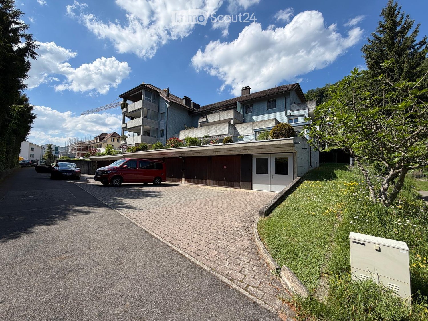 A modern multi-story house with a garage, surrounded by greenery and a paved driveway.