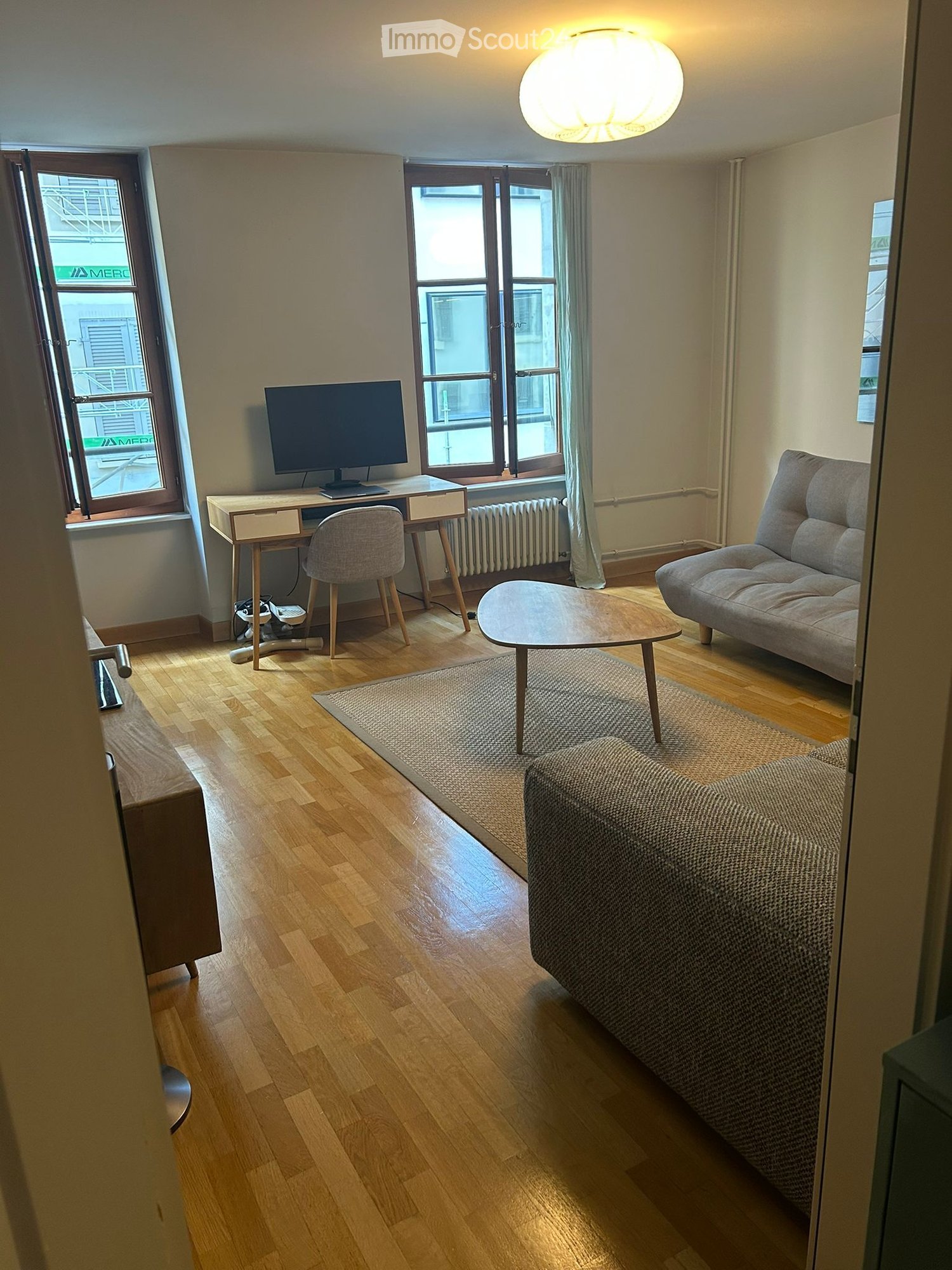 A modern living room with wooden flooring, a sofa, coffee table, desk with a monitor, and a chair.