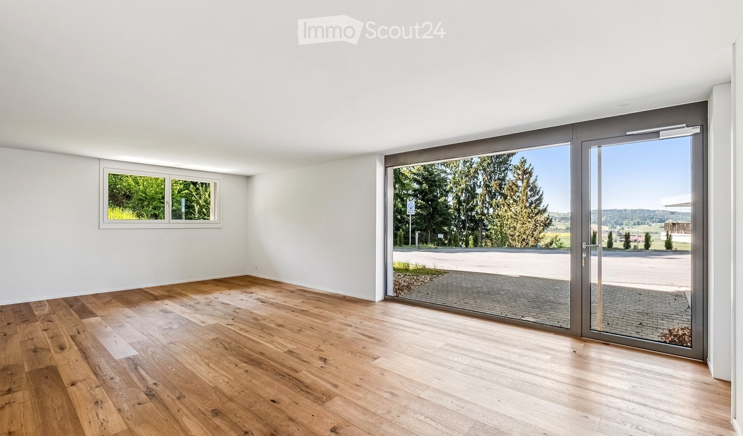 Empty room with wooden flooring, white walls, large sliding glass doors, and a window with a view of greenery.
