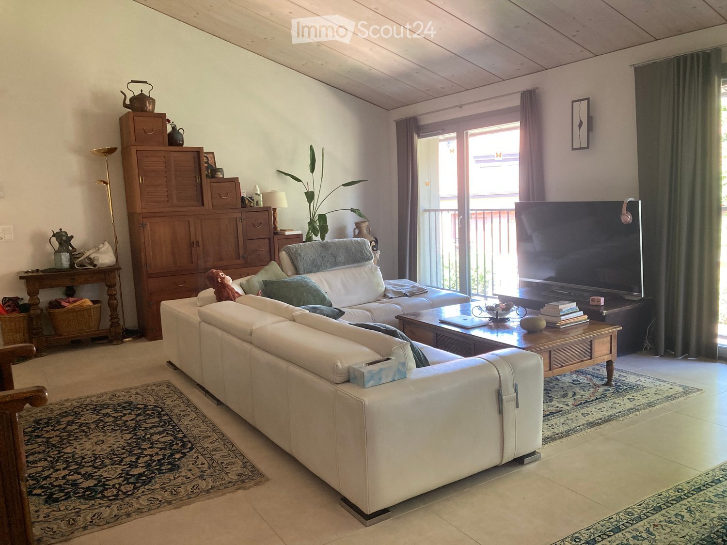 Bright living room with white leather sofa, coffee table, TV, wooden cabinet, and large windows opening to a balcony.