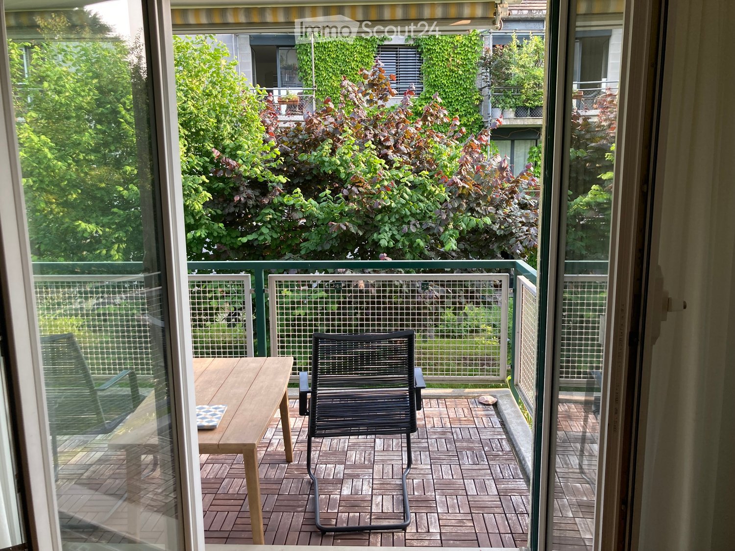 Balcony with wooden table, black chair, and green railing, overlooking a garden with trees and shrubs.
