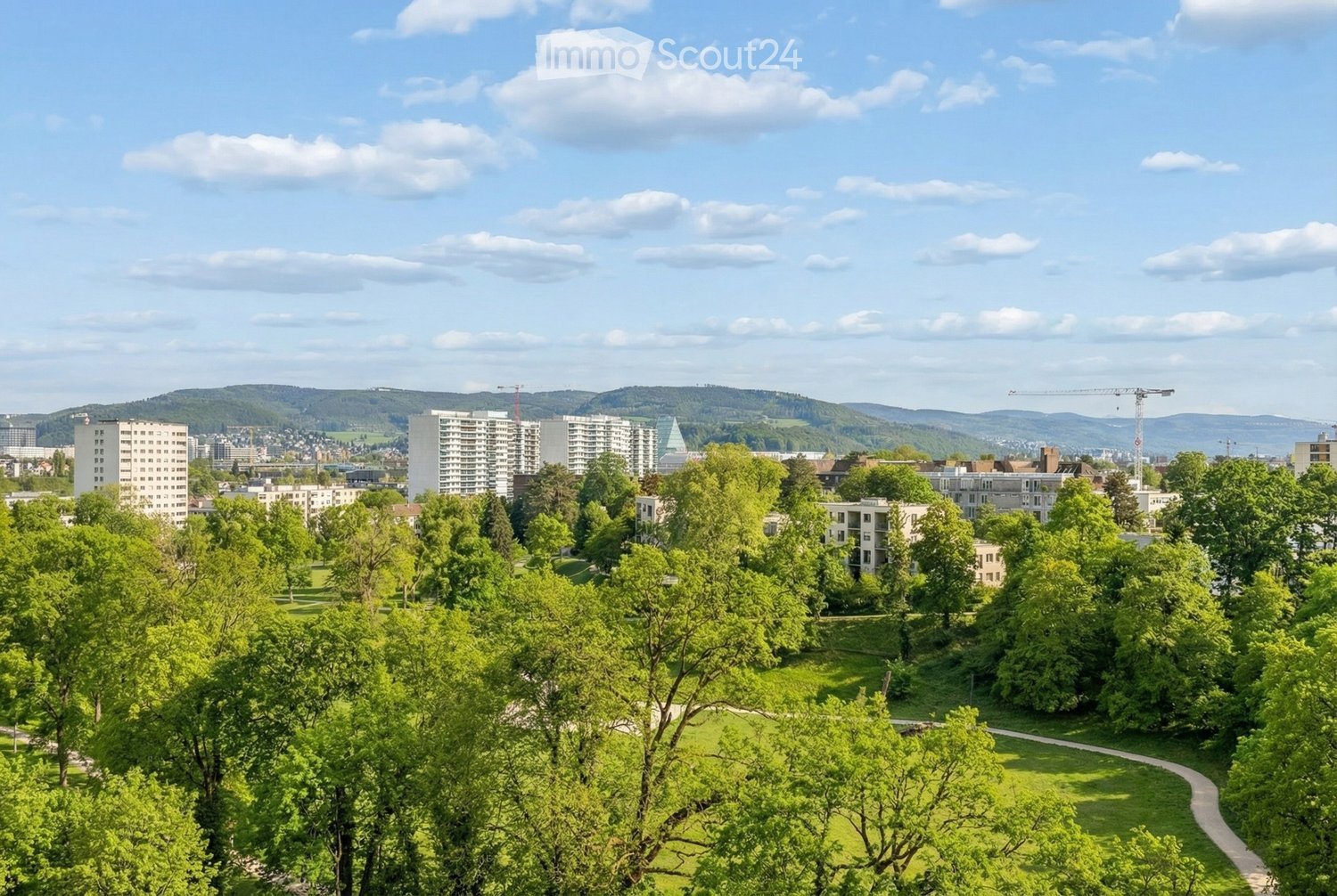 A panoramic view of a cityscape with residential buildings, green spaces, and a distant mountain range under a partly cloudy sky.
