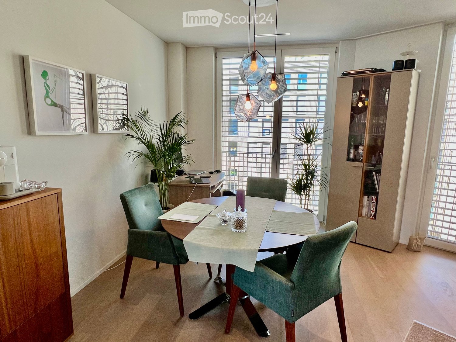 Dining room with a round wooden table, four green upholstered chairs, a cabinet, and a plant near the window.