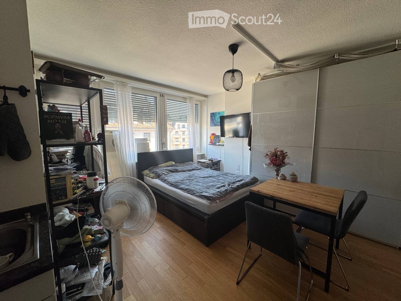 A small, furnished room with a bed, a wooden table with chairs, a fan, a shelf with various items, a mirror, and a television mounted on the wall. The room has wooden flooring and large windows with blinds.