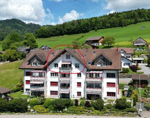 Aerial view of a multi-story residential building with white walls, red shutters, and multiple balconies, surrounded by greenery and other houses in a rural setting.