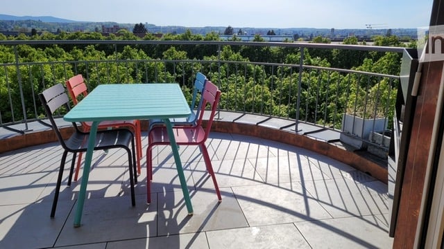 Balcony with a small table and four chairs, overlooking a green landscape with trees and distant cityscape.