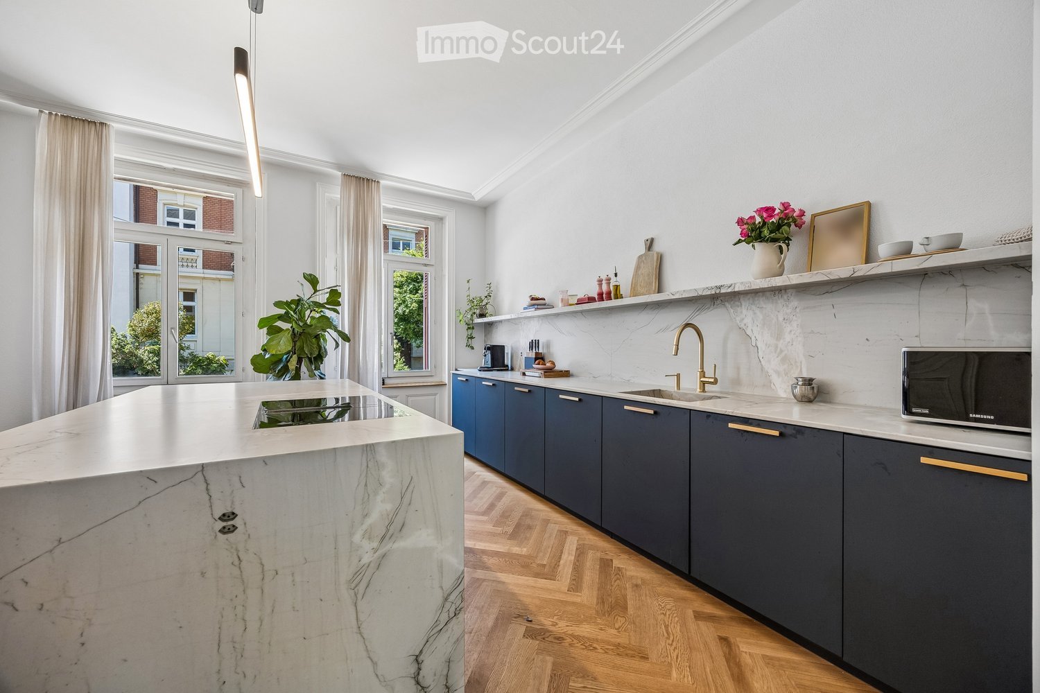 Modern kitchen with marble countertops, dark blue cabinets, a central island, and a sink with a gold faucet. Shelves hold decorative items, and a potted plant adds greenery.