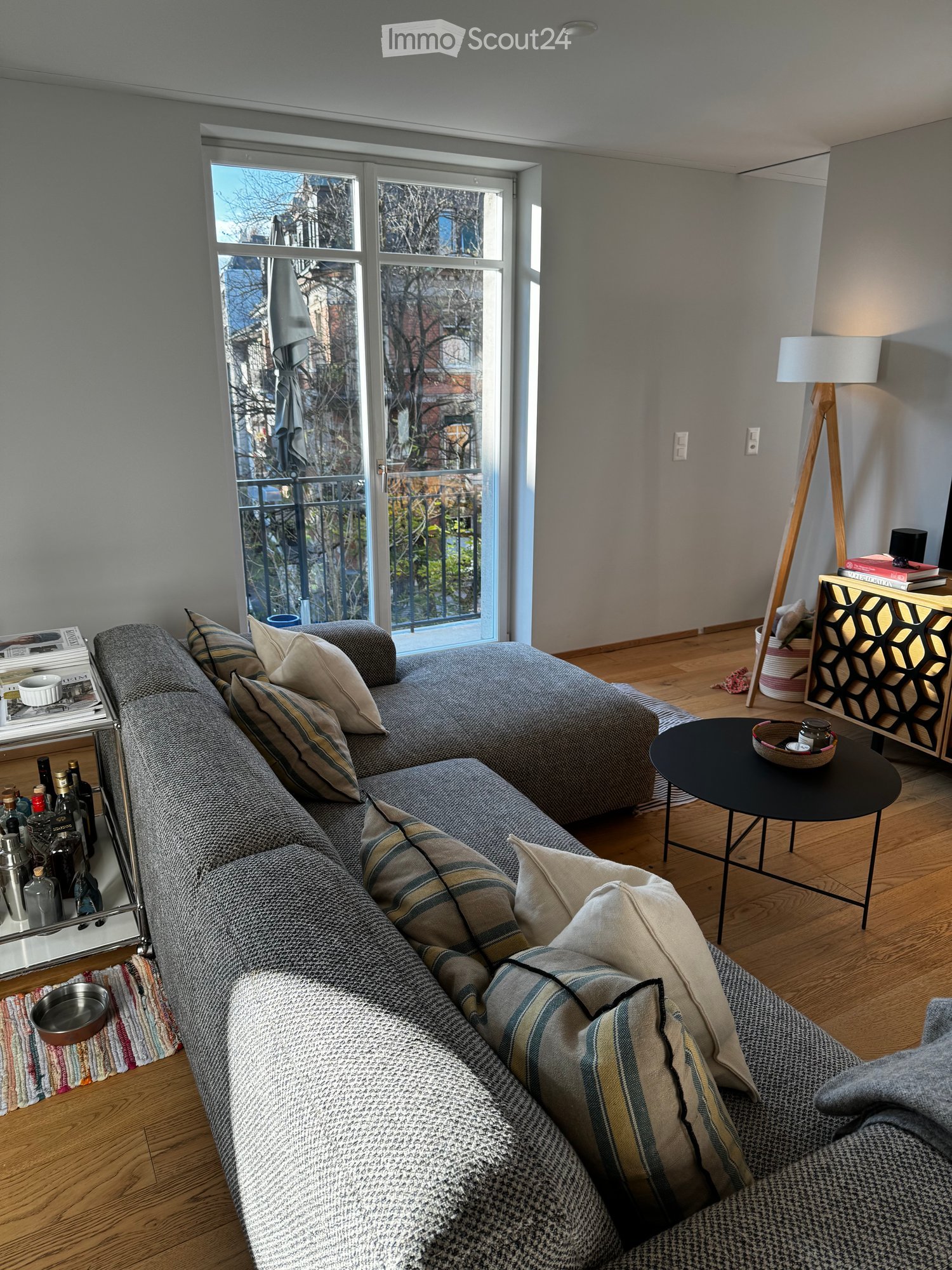 Modern living room with a grey sectional sofa, wooden floor, large window with balcony access, side table, floor lamp, and wine rack.