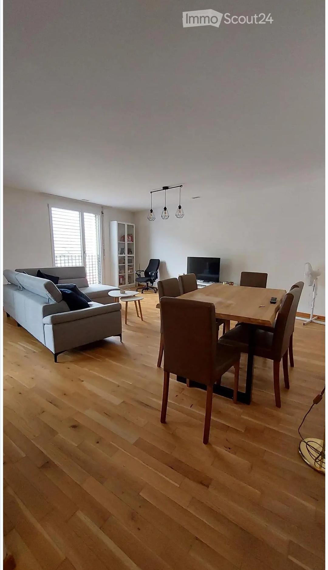A modern, open-plan living and dining area with wooden flooring, a grey sectional sofa, a wooden dining table with chairs, a TV, and a fan. The space is well-lit with pendant lights and has large windows with blinds.